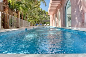 A swimming pool with clear blue water, surrounded by palm trees and a building with arched windows. - The Pink Elephant Hotel (Sant Feliu de Guixols)