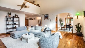 A spacious living area with a large L-shaped sofa, a round coffee table, and a side table. There is a bookshelf with decorative items and a starfish decoration on the wall. A dining area is visible in the background with a table and chairs.