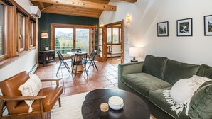 A cozy living area with a green sofa, a brown leather chair, and a round coffee table. There is a dining table with chairs and a view of the outdoors through large windows.