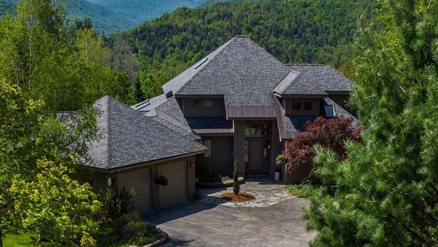 A multi-story house with a stone driveway, surrounded by lush greenery and mountains in the background.