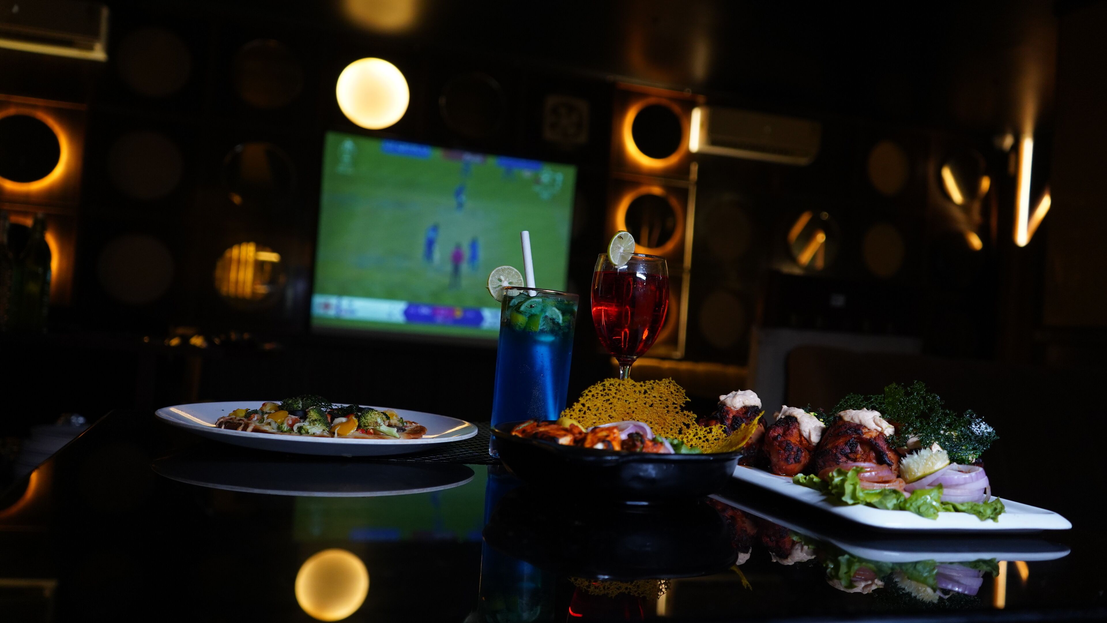 A table with a plate of food, a drink, and a bowl of snacks in a dimly lit room with a TV in the background.