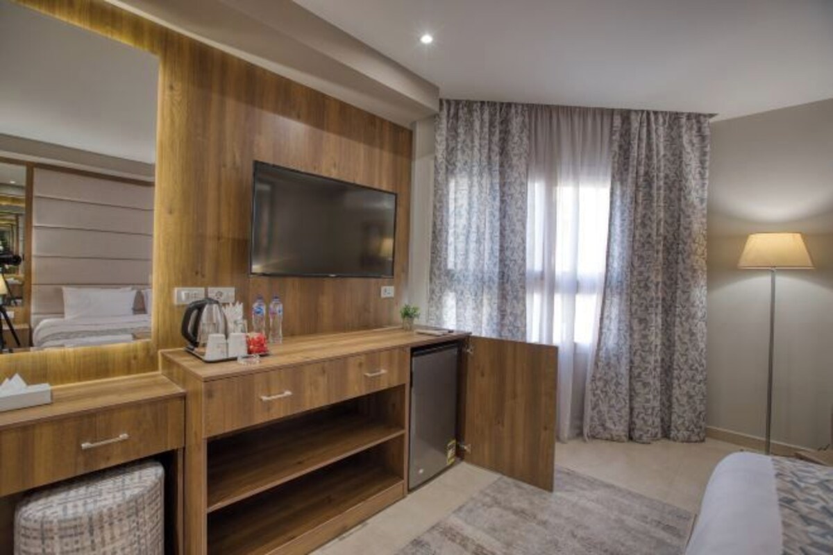 A hotel room with a wooden vanity, a flat-screen TV, a small refrigerator, a bedside table, and a bed.