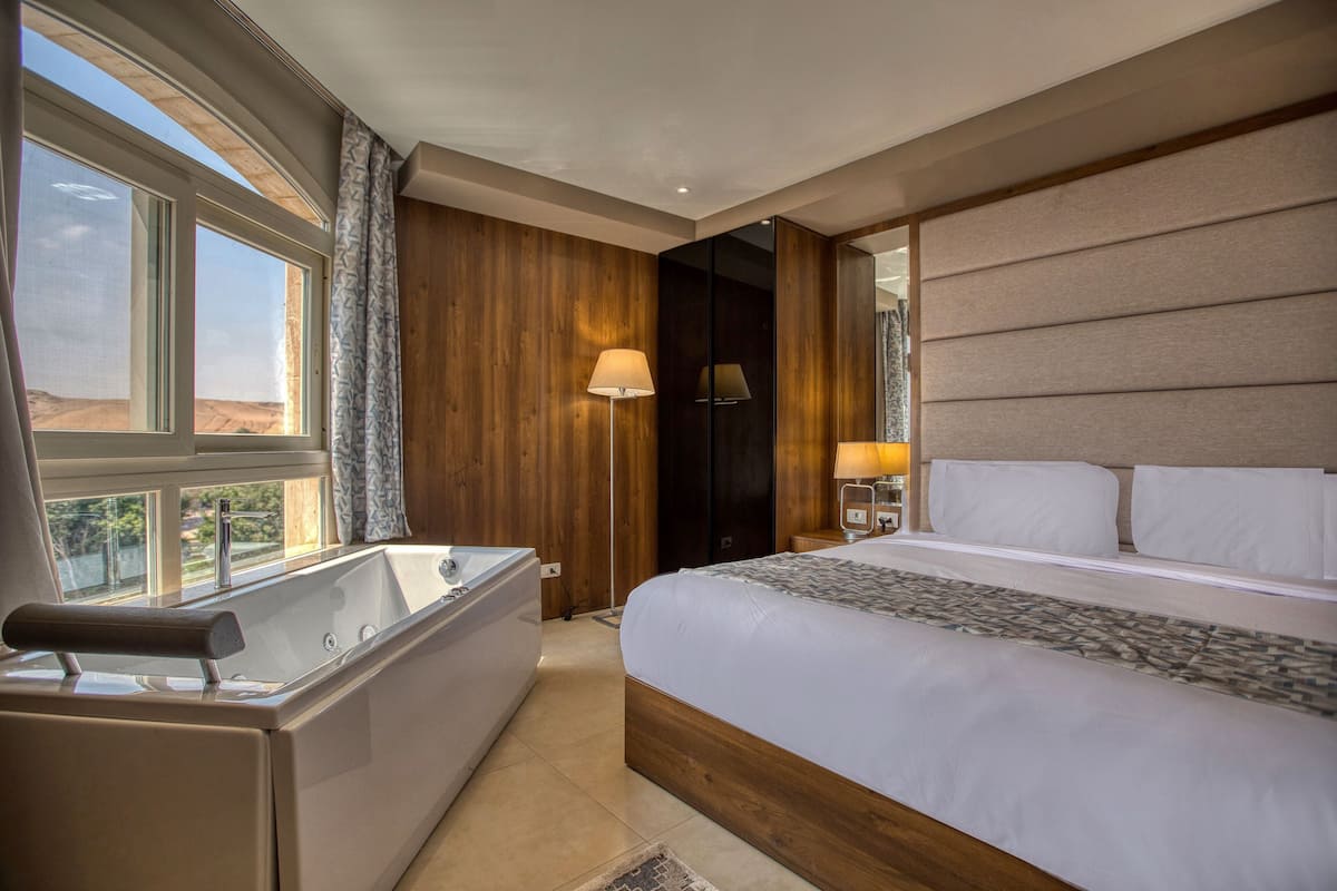 A modern hotel room with a large bed, a separate bathtub, and a large window offering a view of the desert.