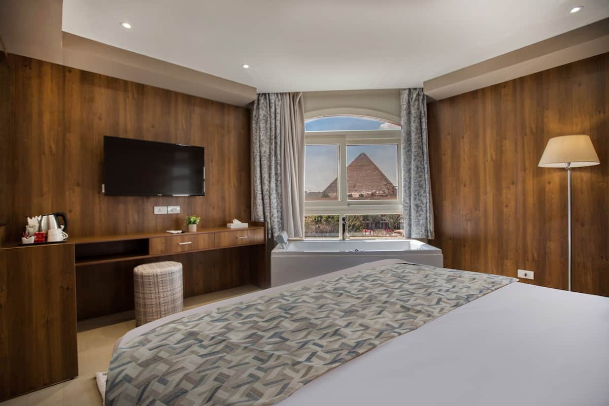 A modern hotel room with a large bed, a flat-screen TV mounted on the wall, a desk with a lamp, and a view of the pyramid.