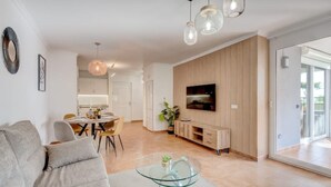 Uma sala de estar moderna com um sofá, uma mesa de centro de vidro, um espaço de refeições com cadeiras, uma cozinha ao fundo e uma TV fixada na parede.