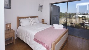Une chambre avec une grande fenêtre, un lit avec une literie blanche, un plaid rose et une tête de lit en bois.
