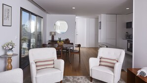 Un salon moderne avec deux fauteuils blancs, une table à manger et un coin cuisine doté de meubles blancs et d’un téléviseur.