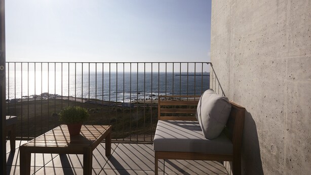 Un balcon avec vue sur l’océan, une chaise en bois, une petite table avec une plante et un mur en béton.