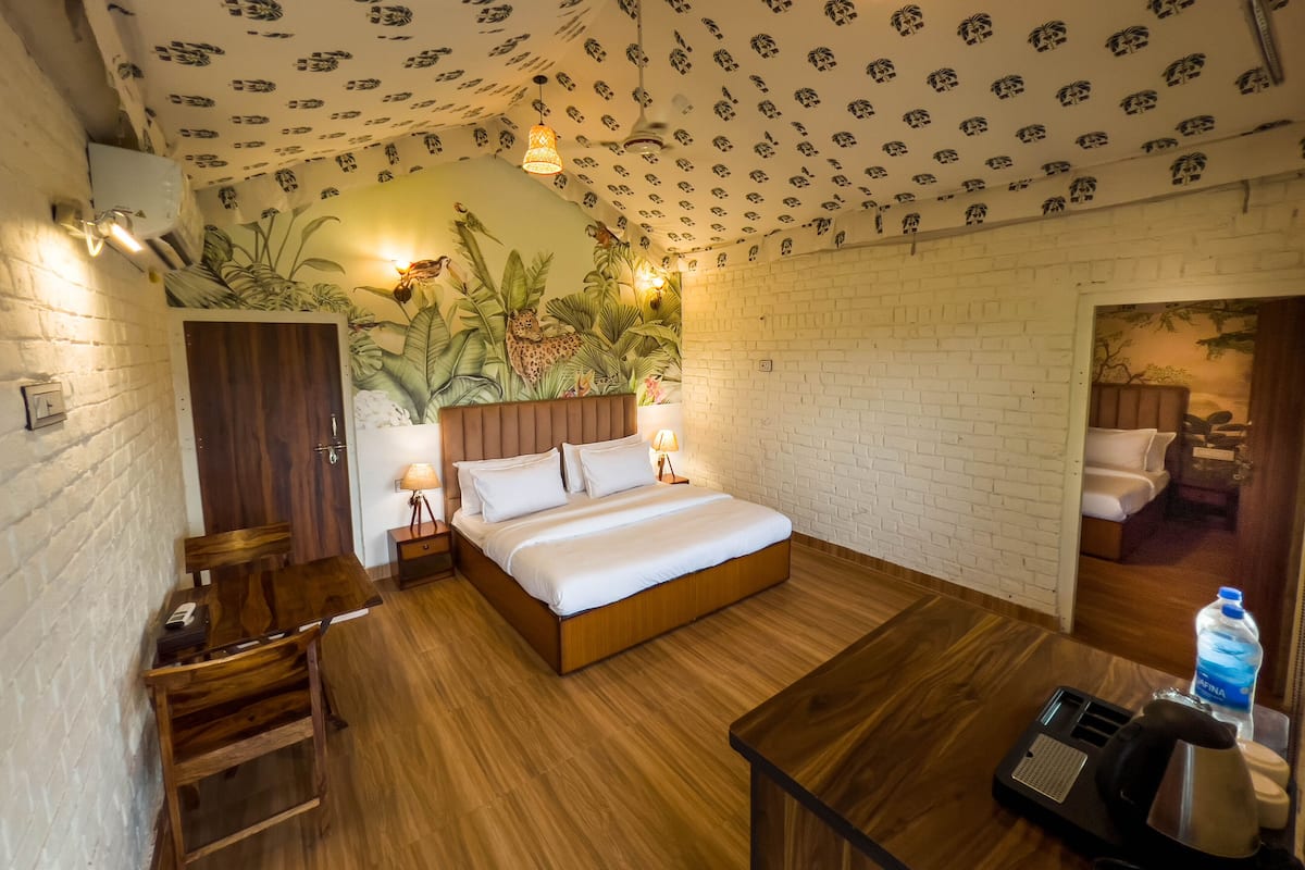 A hotel room with a wooden bed, a brick wall, a mural of tropical plants, a wooden nightstand, and a desk with a kettle and water bottle.
