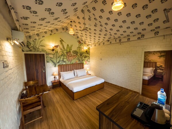 Ein Hotelzimmer mit einem hölzernen Bett, einer Backsteinwand, einem Wandgemälde mit tropischen Pflanzen, einem Nachttisch aus Holz und einem Schreibtisch mit Wasserkocher und Wasserflasche.
