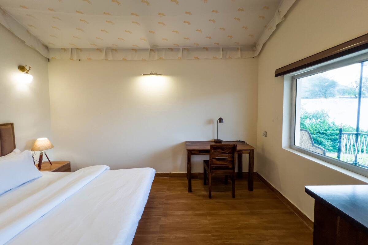A bedroom with a bed, a desk, a chair, a lamp, and a window with a view of greenery.