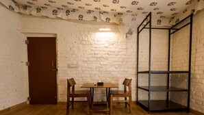 A room with a white brick wall, a wooden table with two chairs, a brown door, and a black metal shelving unit. - Vanbagh Sariska Resort (Alwar)