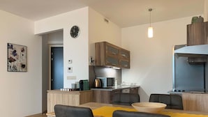 A modern kitchen with wooden cabinets, a built-in microwave, and a kettle. There is a dining area with a wooden table and grey chairs.