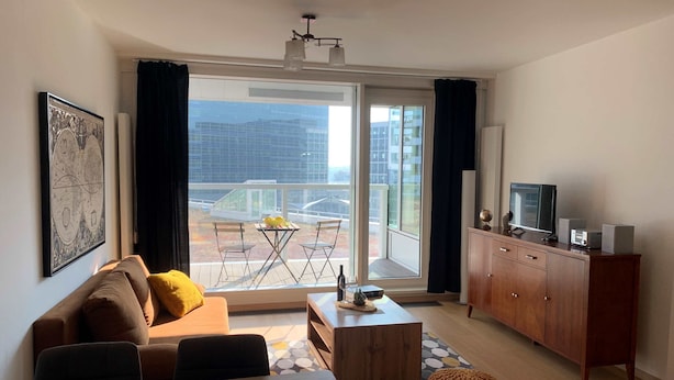 A modern living room with a sofa, a coffee table, a TV, and a balcony with a view of buildings.