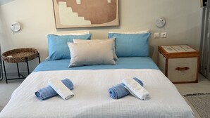 Un lit bien fait, avec des serviettes bleues et blanches, un tableau abstrait accroché au mur, une table de chevet avec une valise, et une petite chaise en osier.