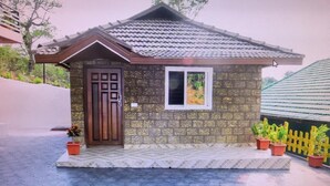 Une petite maison avec un toit en tuiles, une porte en bois et une fenêtre. Des plantes en pot sont disposées devant la maison.