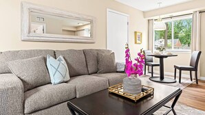 A living room with a grey sofa, a coffee table, a dining table with chairs, a large mirror, and a window with a view of trees and buildings.
