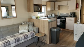 A compact kitchenette with a microwave, stove, and coffee maker. There's a sofa with a plaid blanket and a small side table. - 2 Brightholme 6 Berth With Decking & Enclosed Gard (Burnham-on-Sea)