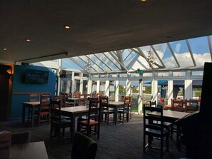 An indoor dining area with tables and chairs, a blue wall with a sign, and a glass ceiling allowing natural light.