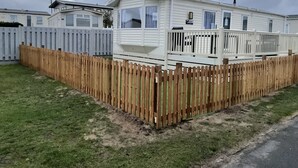 A mobile home with a fenced yard, a white picket fence, and a grassy area. - 2 Brightholme 6 Berth With Decking & Enclosed Gard (Burnham-on-Sea)