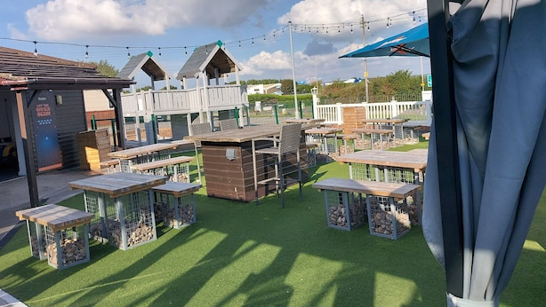 An outdoor seating area with tables and chairs, surrounded by wooden structures and a fenced play area. - 2 Brightholme 6 Berth With Decking & Enclosed Gard (Burnham-on-Sea)
