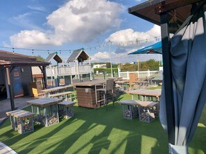 An outdoor seating area with tables and chairs, surrounded by wooden structures and a fenced play area. - 2 Brightholme 6 Berth With Decking & Enclosed Gard (Burnham-on-Sea)