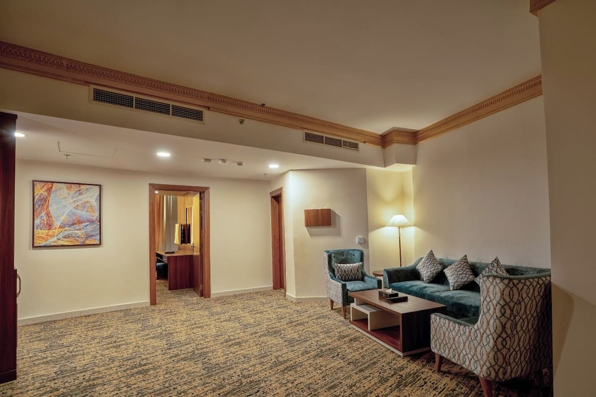 Um hall de hotel com piso em carpete, um conjunto de sofás, uma luminária e uma obra de arte emoldurada na parede.