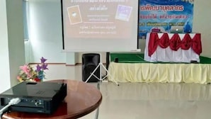 A presentation setup with a projector, screen, and a speaker. There is a table with a tablecloth and a banner in the background. - Ruen Bua resident (Warin Chamrap)