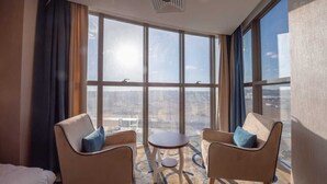 A hotel room with a large window offering a city view, two armchairs, a small round table, and a bed.