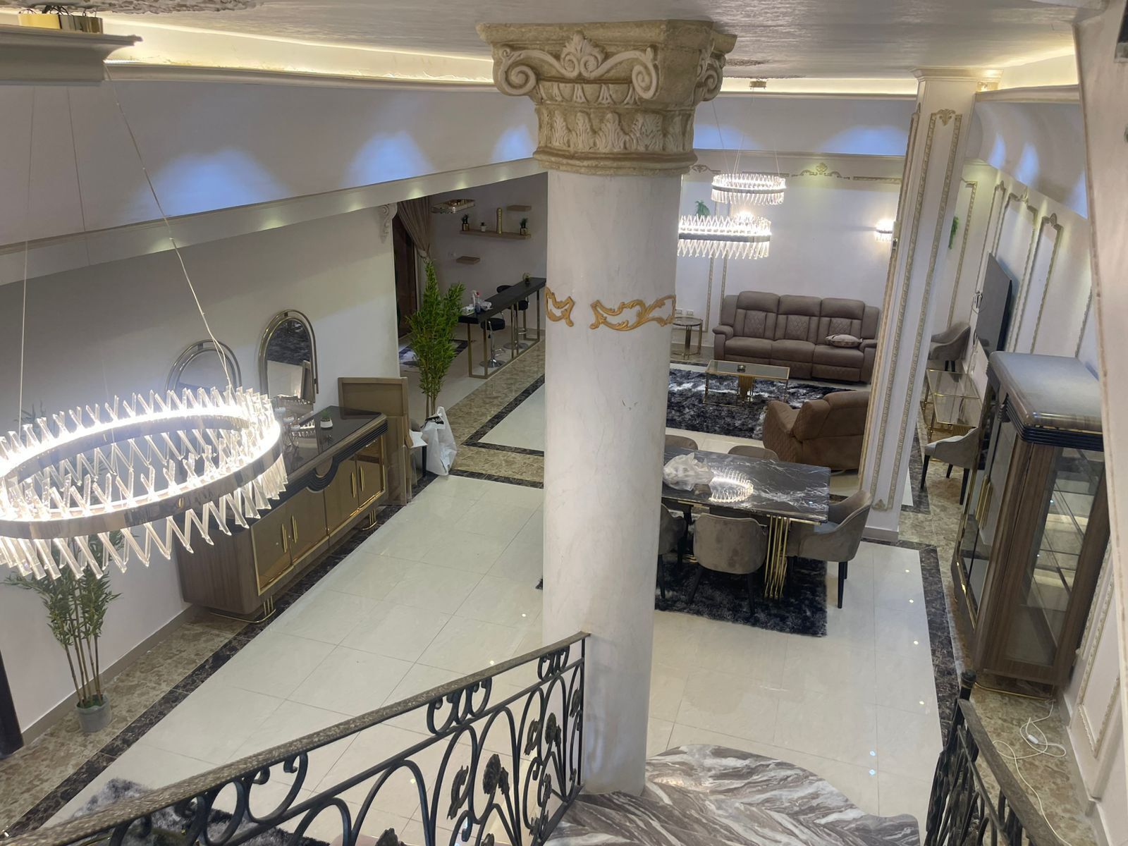A spacious lobby with a high ceiling, a central column, a chandelier, a seating area with sofas, and a dining table.