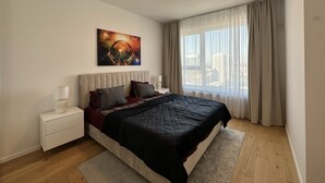 Een moderne slaapkamer met een groot bed, een nachtkastje, een raam met gordijnen en uitzicht op een stadsgezicht.