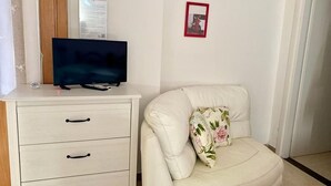 A room with a white sofa, a small white cabinet with drawers, a flat-screen TV on top, a framed picture on the wall, and a door in the background. - B&B La cascata (Noci)