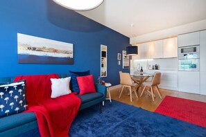 Un soggiorno moderno con un divano blu, cuscini rossi e bianchi, e un tappeto blu. C'è una zona pranzo con sedie in legno e un tavolo, e una cucina con mobili e elettrodomestici bianchi.