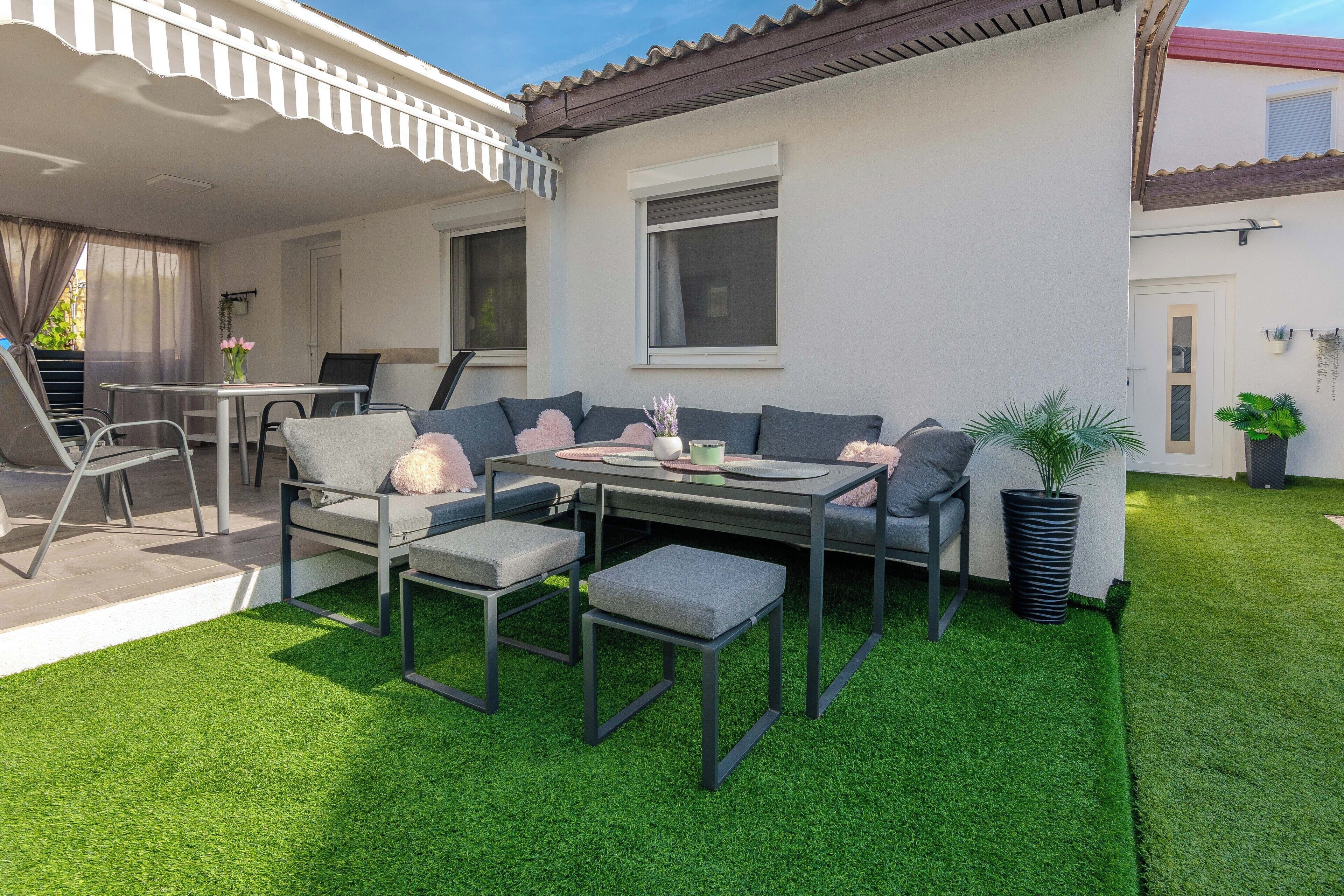 A patio with a table and chairs, surrounded by artificial grass, and a potted plant.