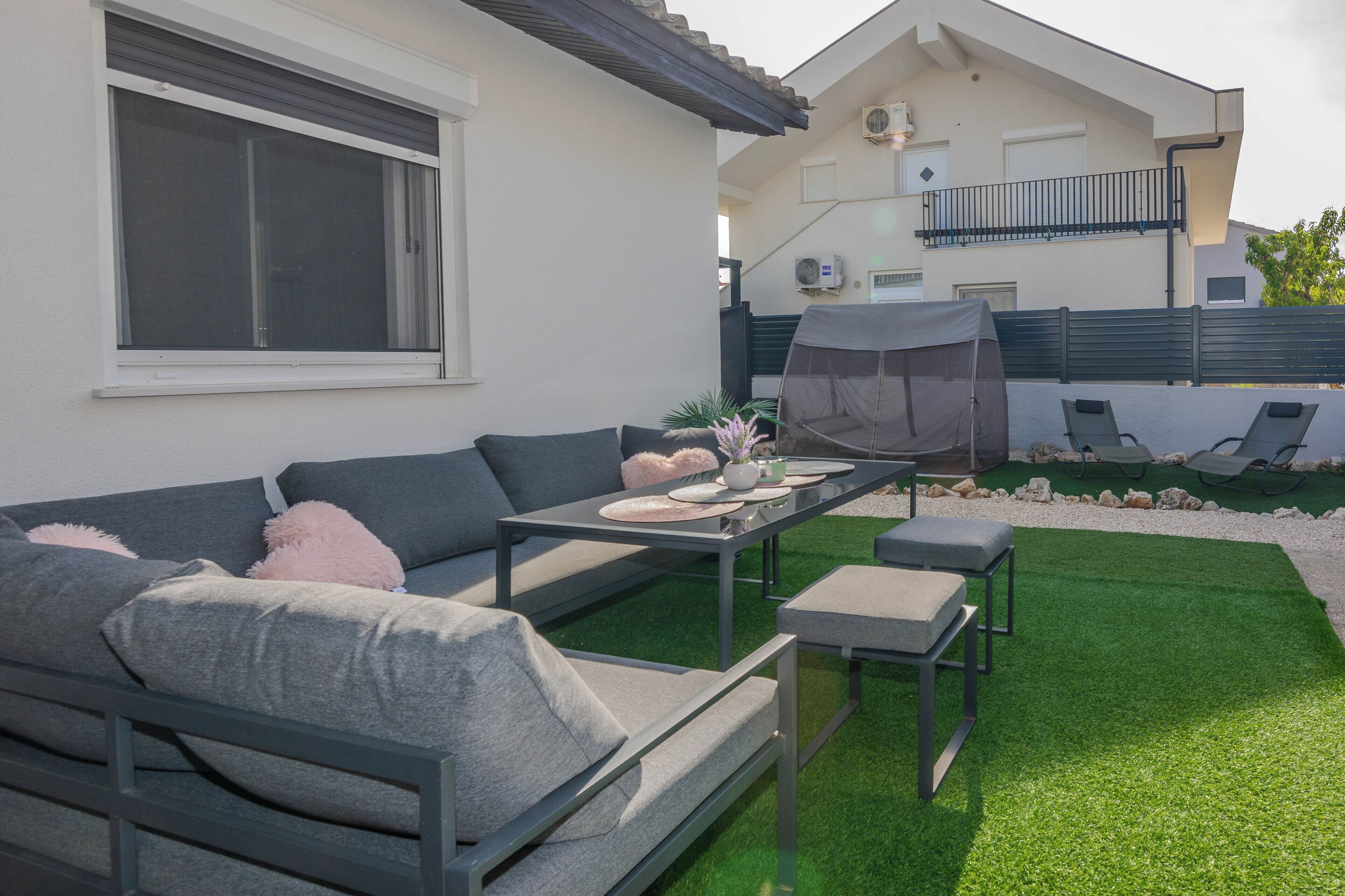 A modern outdoor seating area with a sofa, table, and chairs, surrounded by a well-maintained lawn and a house in the background.