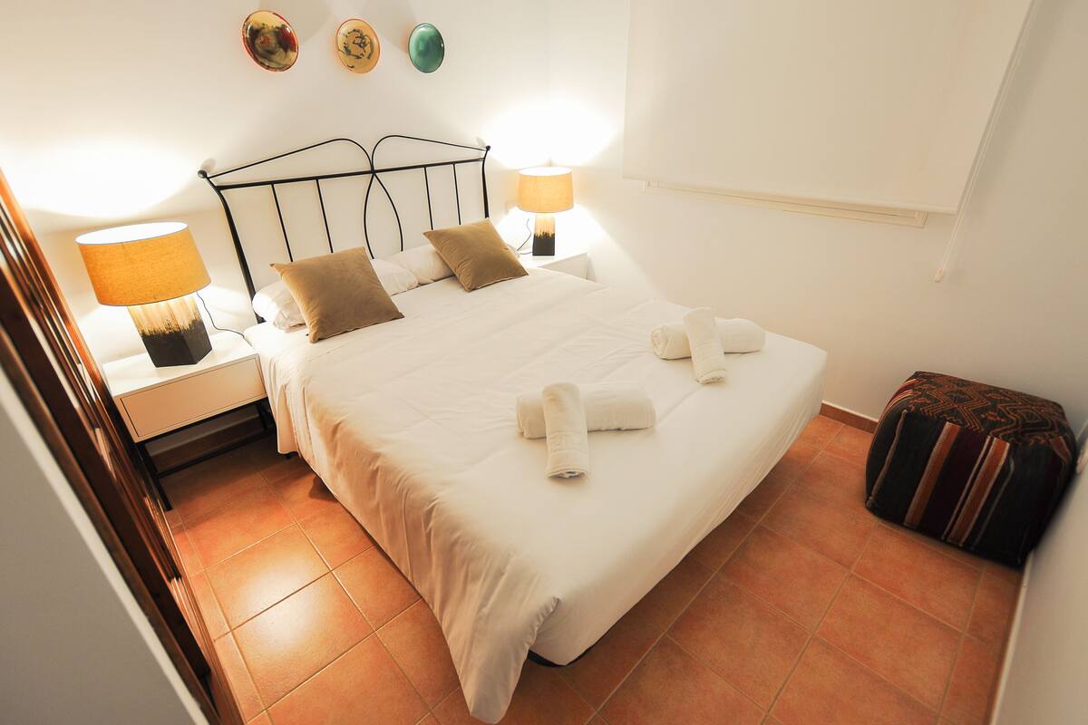 Una cama bien hecha con sábanas blancas y dos almohadas, una mesita de noche con una lámpara y un pequeño otoman con un cubre lecho estampado.