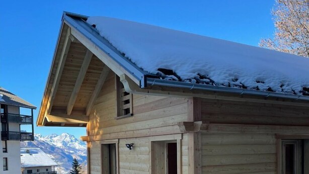 Eine Holzhütte mit Balkon, einem mit Schnee bedeckten Dach und Blick auf verschneite Berge im Hintergrund.