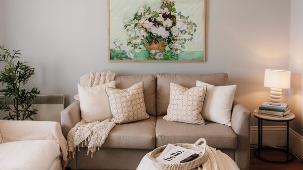 Una sala de estar con un sofá color beige, un reposapiés blanco, una lámpara colgante, un cuadro floral enmarco y una mesita de noche con una lámpara.