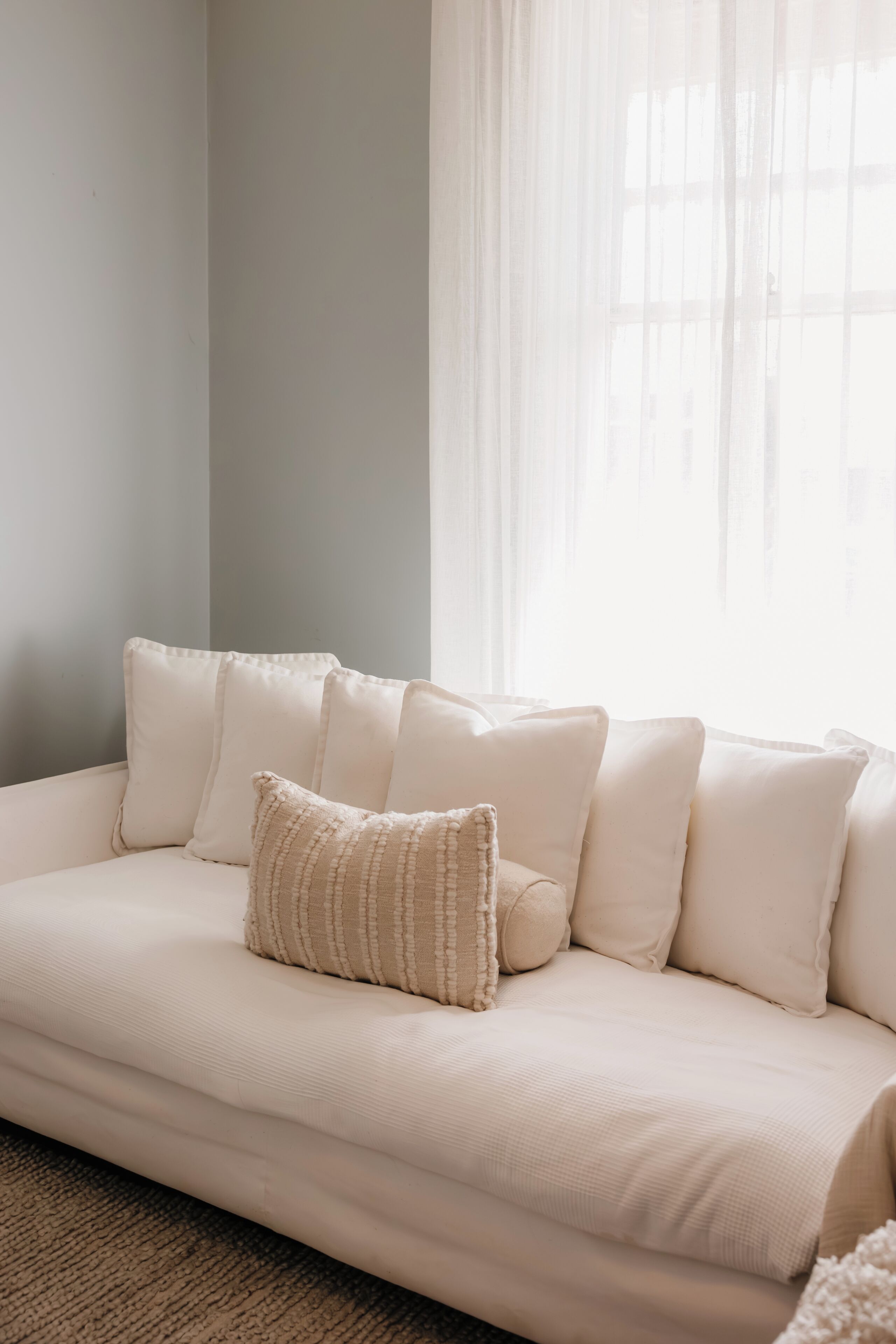A beige sofa with multiple cushions in a room with a plain wall and a window with sheer curtains.
