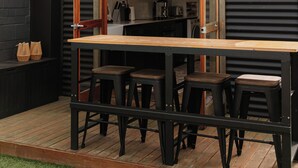 A patio with a wooden table and stools, a black metal wall, and a brick building in the background.