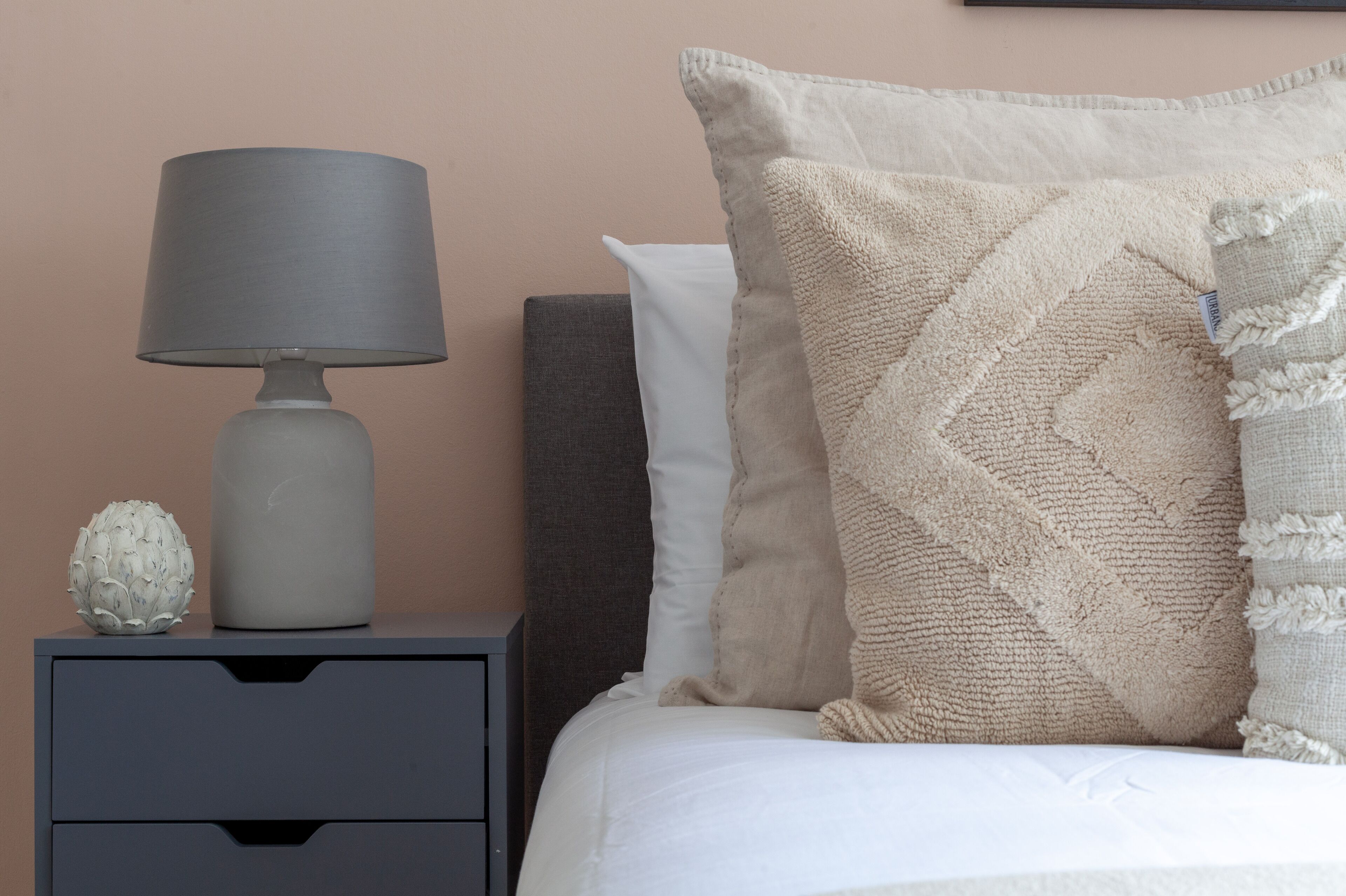 A neatly made bed with a white pillow and a textured beige pillow, a grey lamp, and a decorative sphere on a grey nightstand.