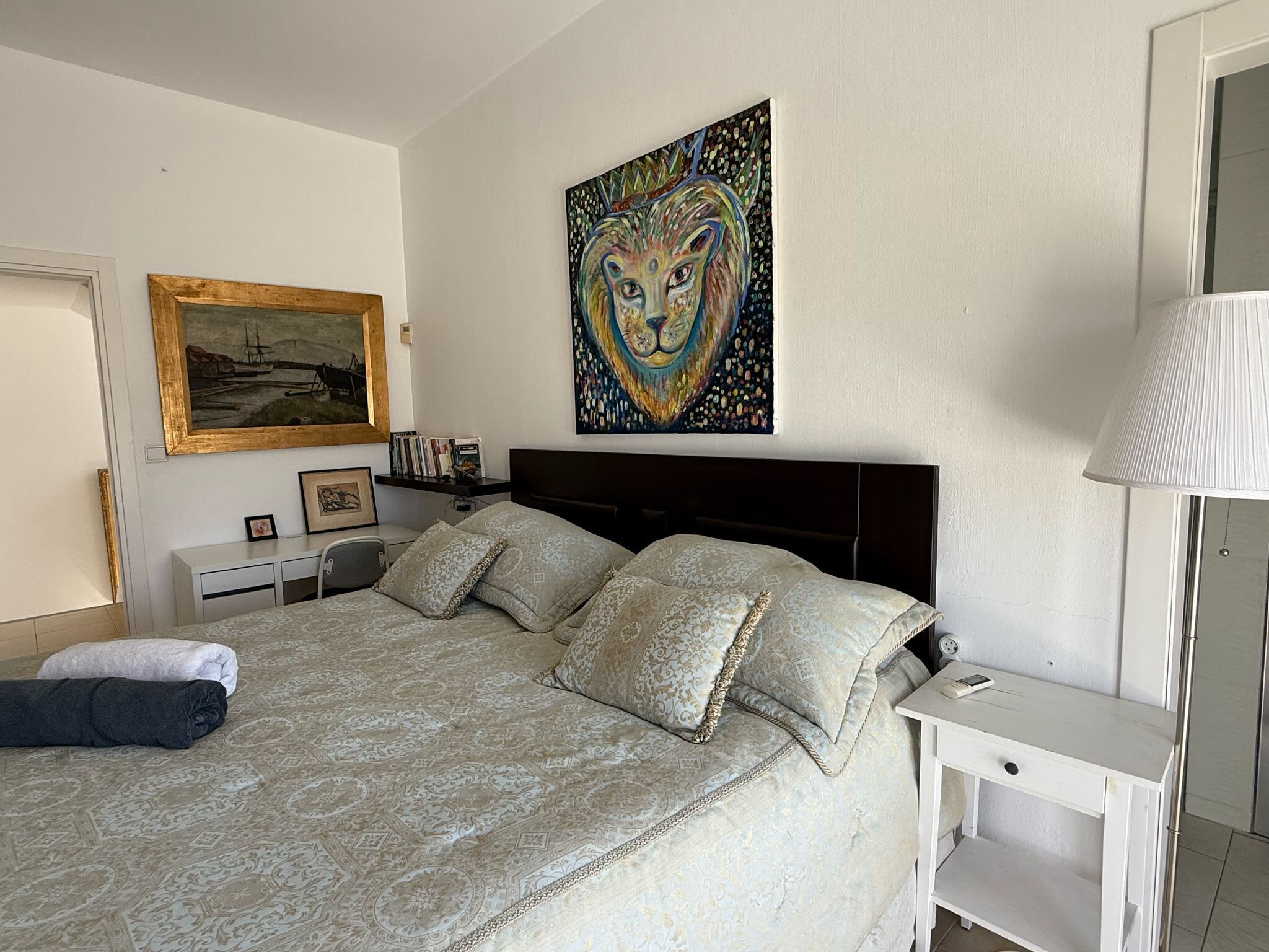 Ein Schlafzimmer mit einem Bett, einem Nachttisch, einer Lampe, einem Gemälde und einem gerahmten Bild an der Wand.