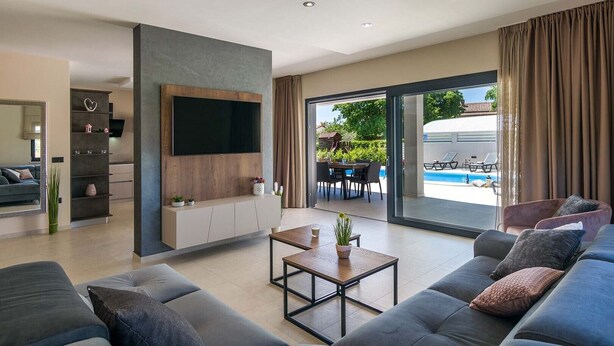 Un soggiorno moderno con una grande TV, un divano, un tavolino e vista su un’area piscina esterna.