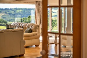 Relax in a spacious living room with a plush sofa facing a scenic view of rolling hills and lush greenery.