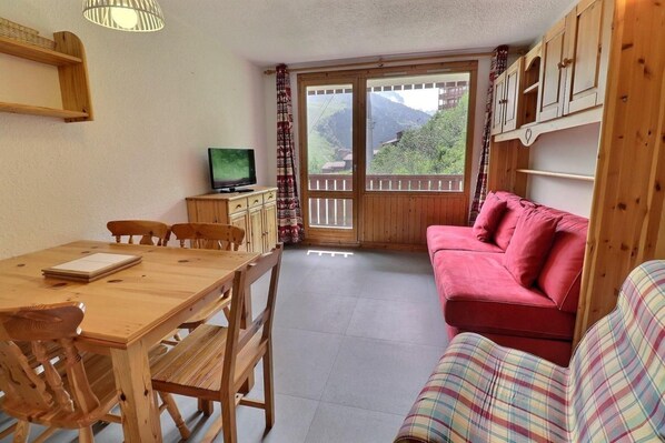 A wooden dining table with chairs, a red sofa, a television on a wooden cabinet, and a view of mountains through a glass door. - 250m from ski slopes, 4th floor, balcony, 26m², Méribel-Mottaret (Méribel - Les Allues)