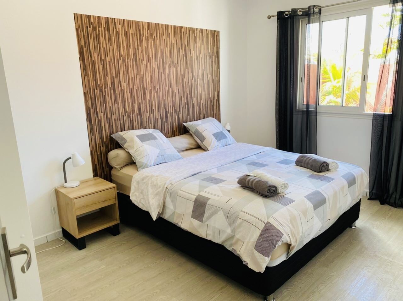 Ein ordentlich bezogenes Bett mit einem geometrisch gemusterten Überwurf, einer Nachttischlampe und einem Holz-Kopfteil.
