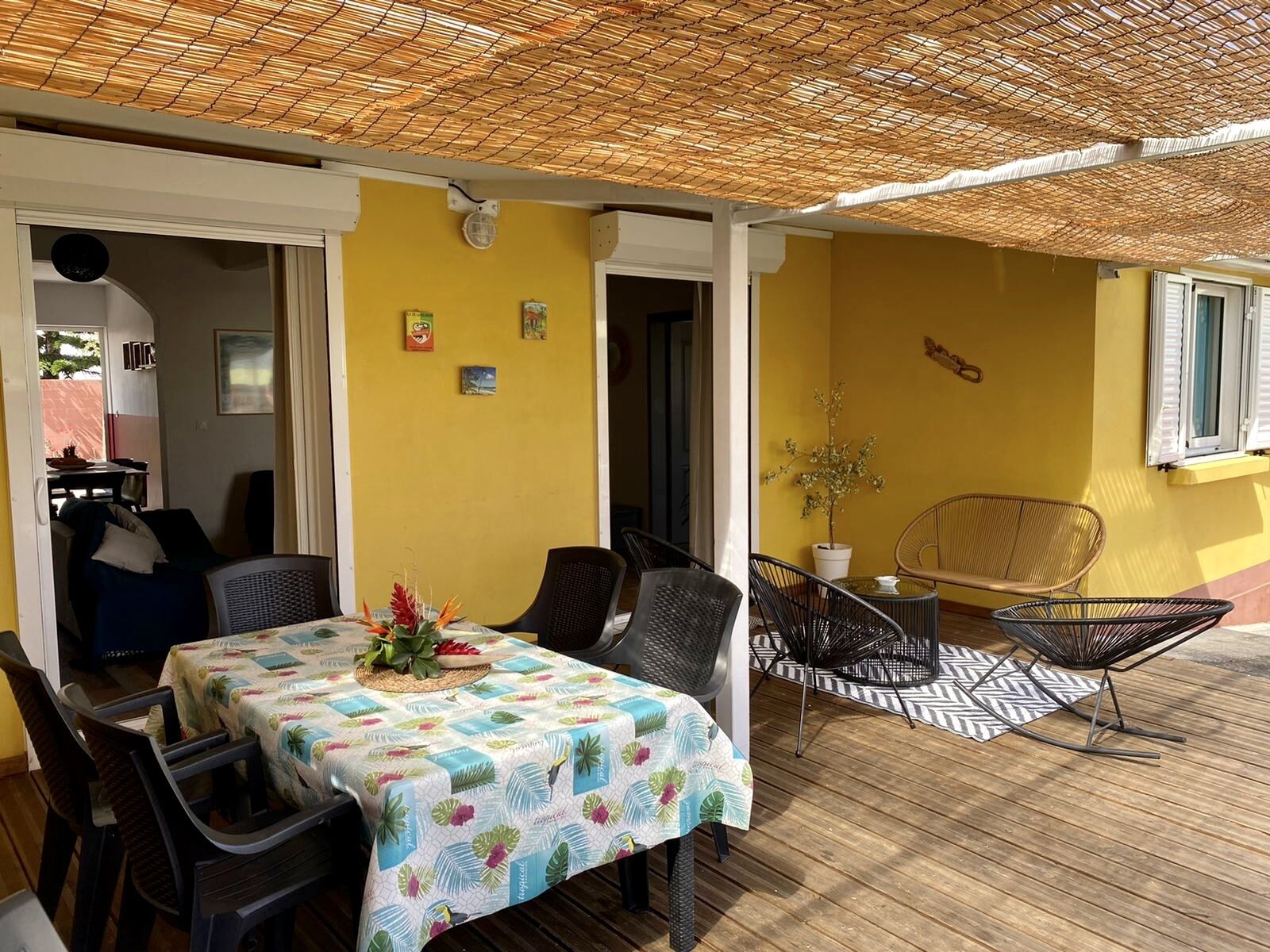 An outdoor dining area with a table covered in a floral tablecloth, surrounded by chairs, under a thatched roof.