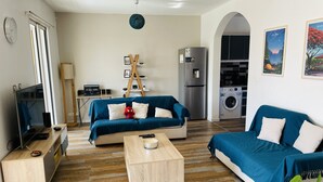 Una sala de estar moderna con dos sofás azules, una mesa de centro, un televisor y una cocina con refrigerador y lavadora.