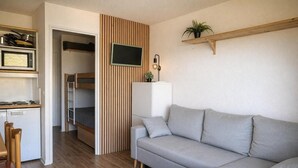 Un espacio habitable compacto con kitchenette, un sofá gris, un televisor montado en un panel de madera y una pequeña planta en un estante.