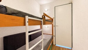 Una cama litera con cabecera de madera y una escalera blanca, una puerta blanca y una ventana pequeña con una lámpara.